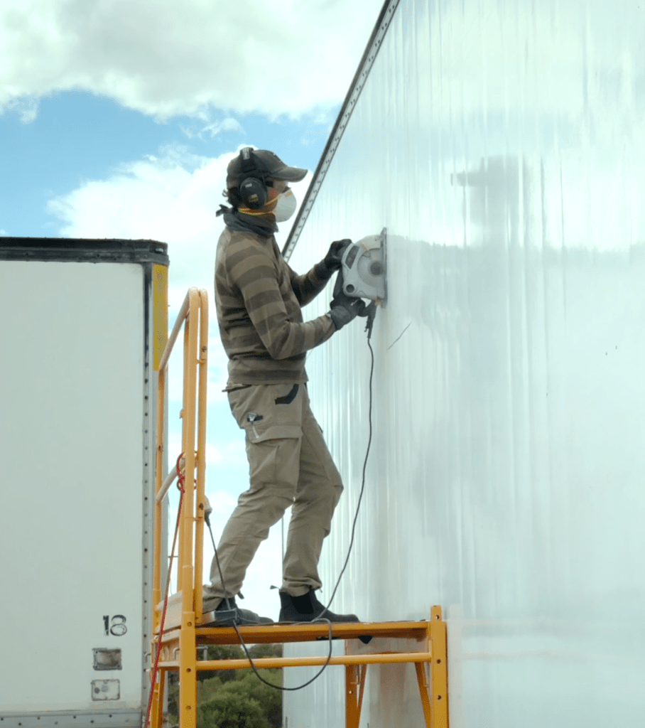 A person using a circular saw to cut the exterior wall of a semi-trailer while standing on a yellow ladder, wearing safety gear including headphones and a dust mask, against a backdrop of partly cloudy blue sky.