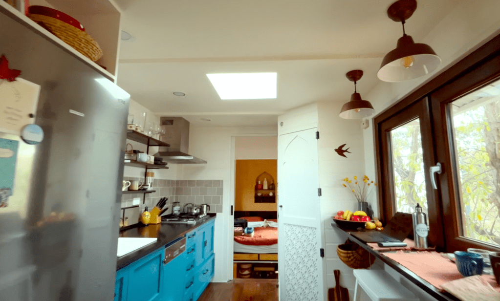 Interior view of a tiny house built inside a semi-trailer, featuring a kitchen area with blue cabinets, a refrigerator, shelves with dishes, and large windows allowing natural light.