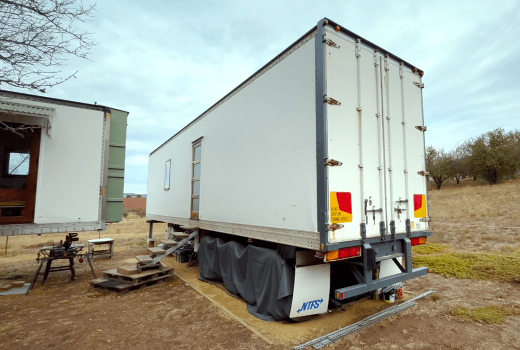 An ex-refrigerated semi-trailer converted into a tiny house, showcasing its rear view with closed doors and a simple wooden staircase leading up to its entrance.